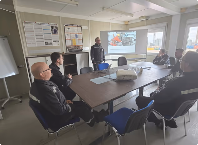 Sechs Männer in Arbeitskleidung sitzen um einen Konferenztisch, während ein Mann vor einer Leinwand eine Präsentation hält.