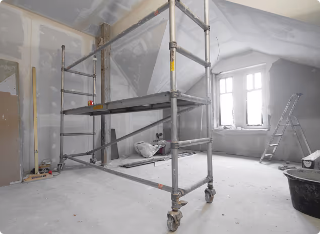 Innenraum einer Baustelle mit Gerüst, Leiter und unvollständig verputzten Wänden unter einem Dachfenster.