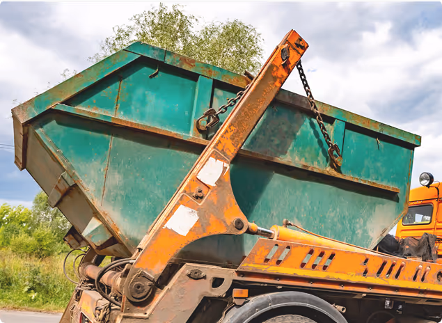 Abfallcontainer wird von einem orangefarbenen LKW mit Hydraulikarm transportiert, bewölkter Himmel im Hintergrund.