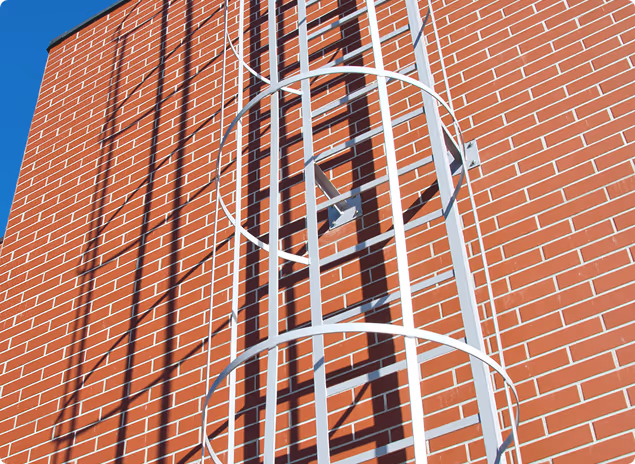 Weiße Metallleiter mit Schutzkäfig an einer roten Backsteinwand bei sonnigem Himmel.