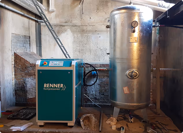 Blauer Renner Kompressor neben einem großen silbernen Druckbehälter in einem industriellen Raum mit Betonwänden und Rohren.
