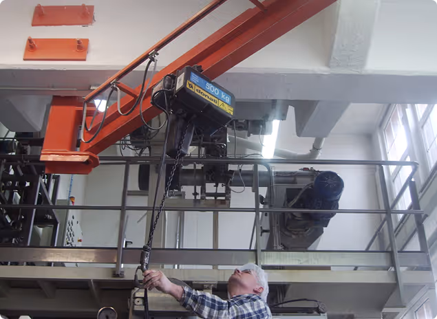 Älterer Mann bedient einen 500-Kilogramm-Kran in einer industriellen Werkstatt mit Metallstruktur und großen Fenstern.