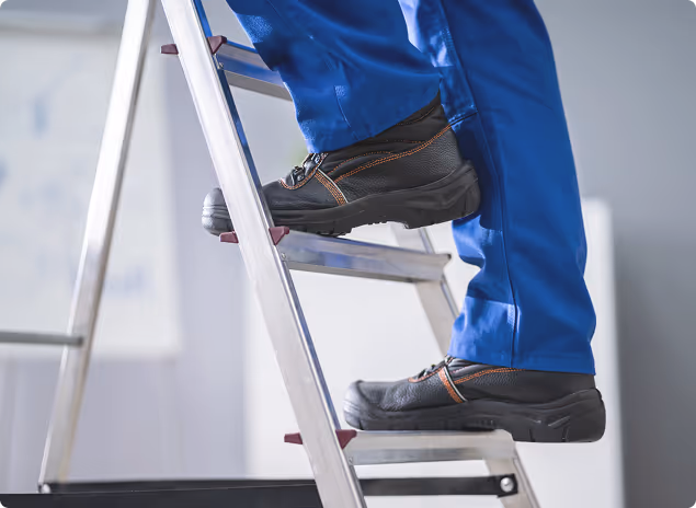 Person mit blauer Arbeitskleidung und schwarzen Sicherheitsschuhen steigt auf eine Aluminiumleiter.