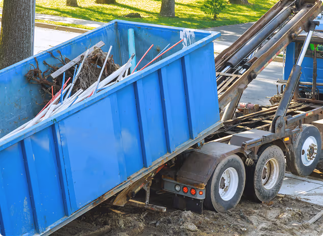 Blauer Müllcontainer auf einem LKW, gefüllt mit Bauabfällen wie Rohren und Ästen, neben einer Straße mit Rasen und Bäumen.