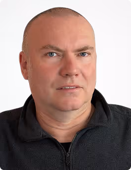 Nahaufnahme eines Mannes mit kurzem Haar, der einen dunklen Reißverschluss-Pullover trägt, vor hellem Hintergrund.