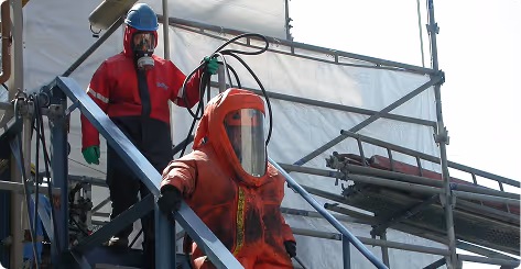 Zwei Personen in Schutzanzügen und Atemmasken arbeiten auf einer Baustelle mit Gerüst.